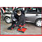 A man in work clothes repairs a car, sitting on a red rolling stool.