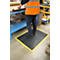 A worker stands on a black work mat with a yellow border. He wears an orange vest and works at a table.