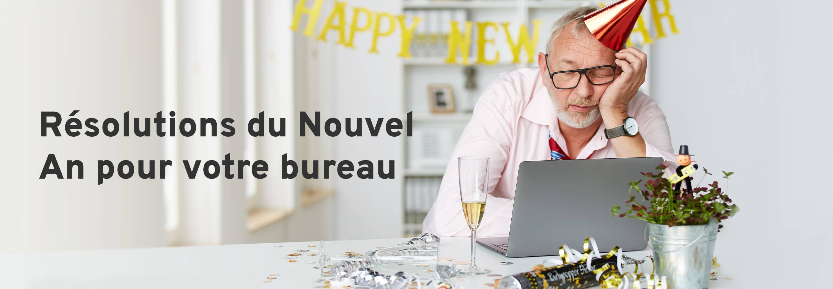 Un homme au chapeau de fête somnole devant un ordinateur portable. Un verre de champagne est posé sur son bureau et la table est décorée pour le Nouvel An. 