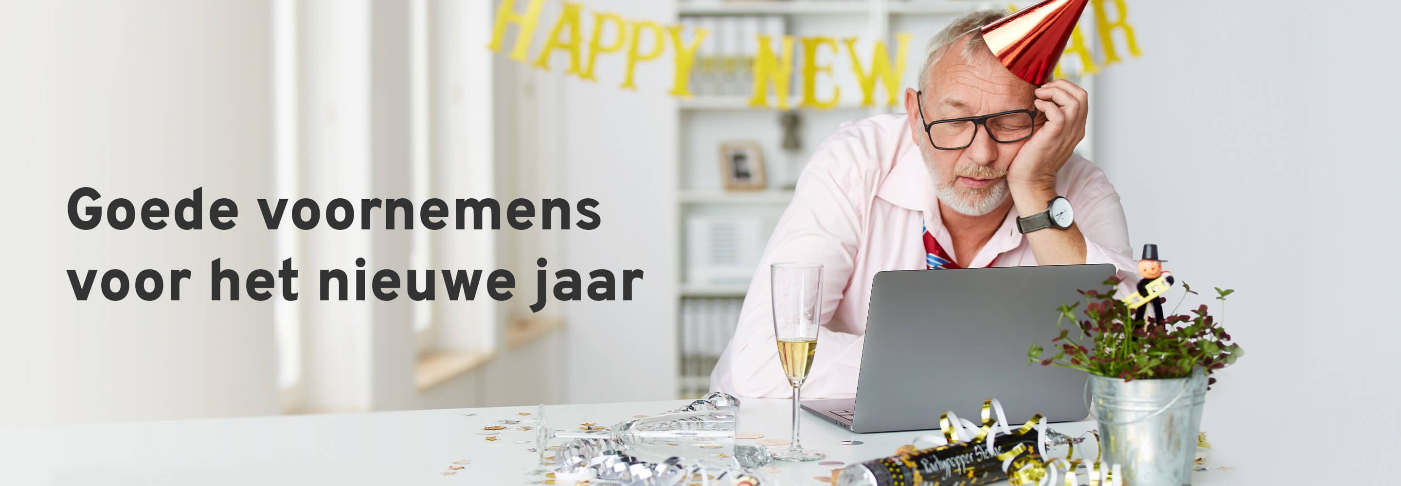 Un homme avec un chapeau de fête somnole devant un ordinateur portable. Un verre de champagne est posé sur son bureau et la table est décorée pour le Nouvel An. 