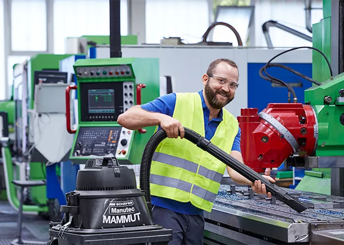 Un employé aspire une machine à l'aide d'un aspirateur industriel tout en souriant à la caméra.
