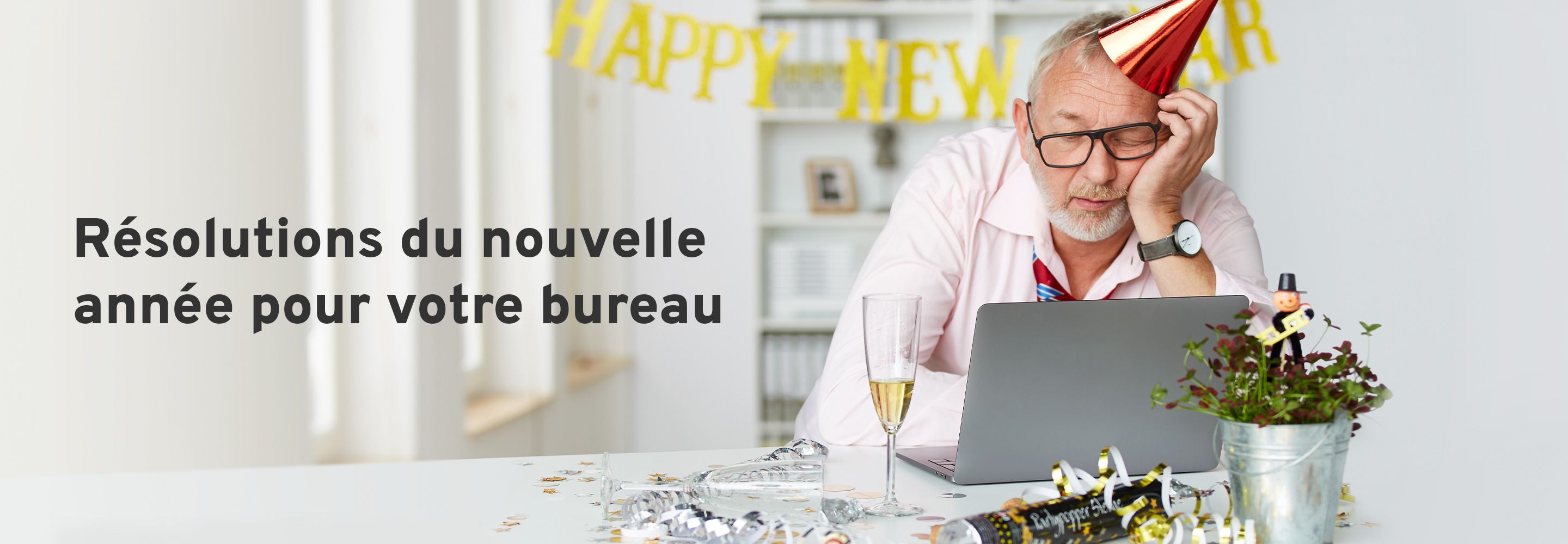 Un homme au chapeau de fête somnole devant un ordinateur portable. Un verre de champagne est posé sur son bureau et la table est décorée pour le Nouvel An. 