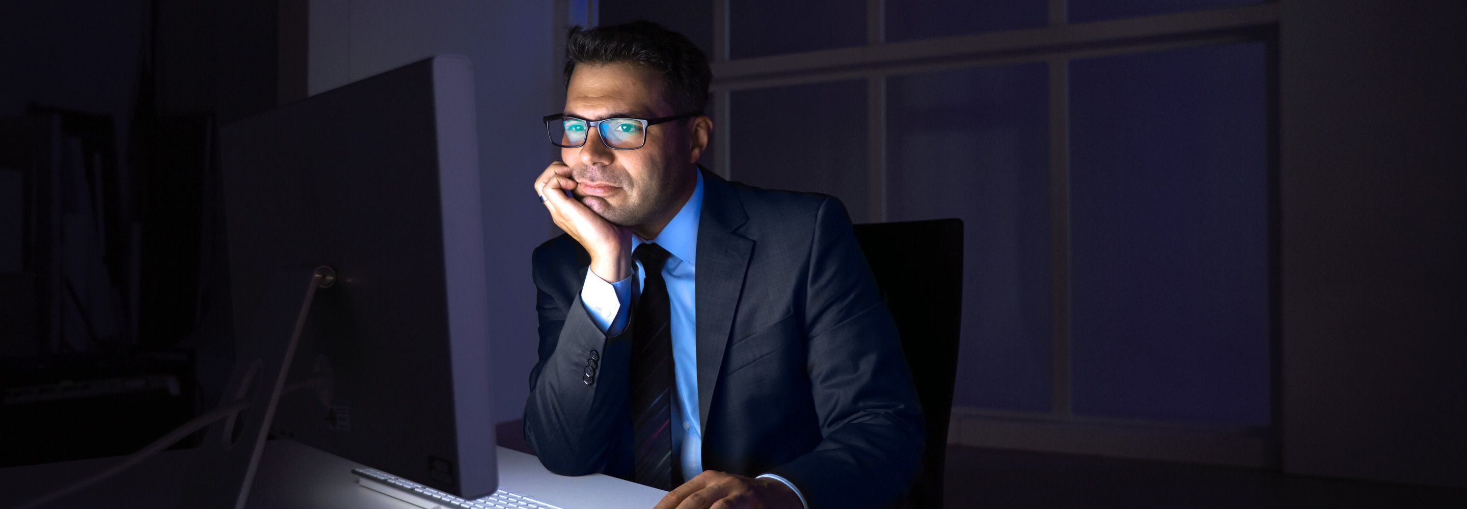 Een man in pak zit in een donker kantoor dat alleen wordt verlicht door het scherm waaraan hij werkt.