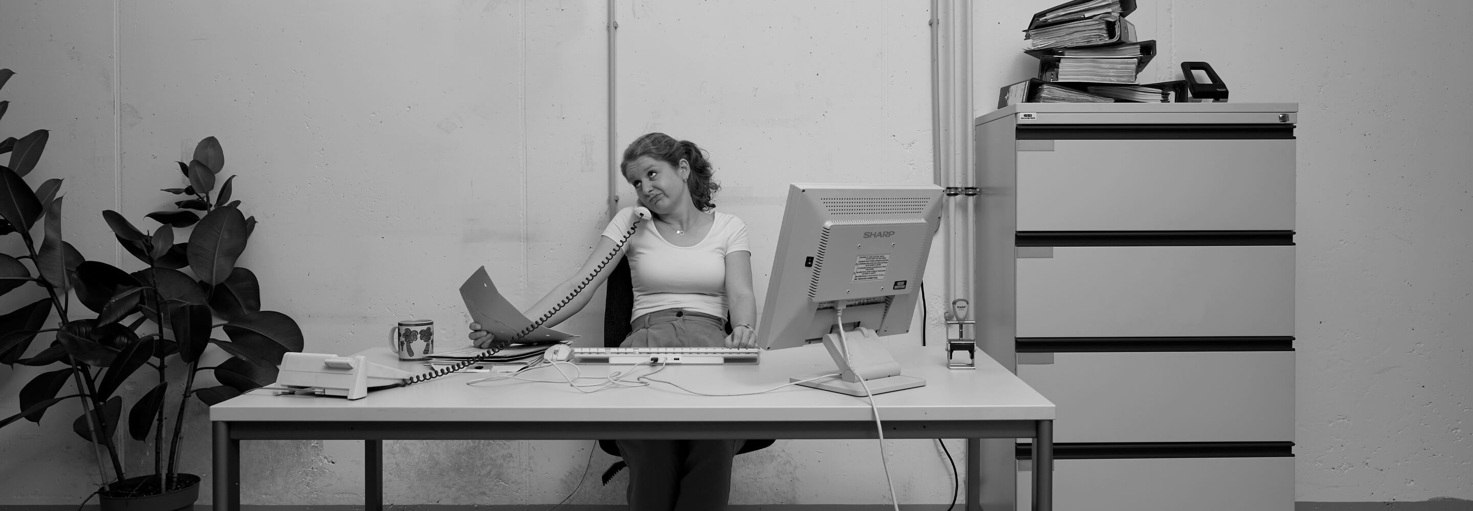 Een vrouw zit aan een bureau aan de telefoon en vervormt haar gezicht. De foto is in zwart-wit.
