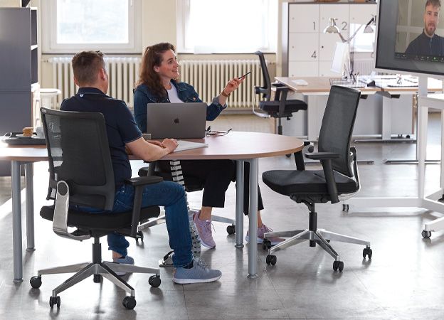Konferenzraum mit Bürostühlen und Whiteboard, in dem Mitarbeiterinnen und Mitarbeiter ein hybrides Meeting abhalten.