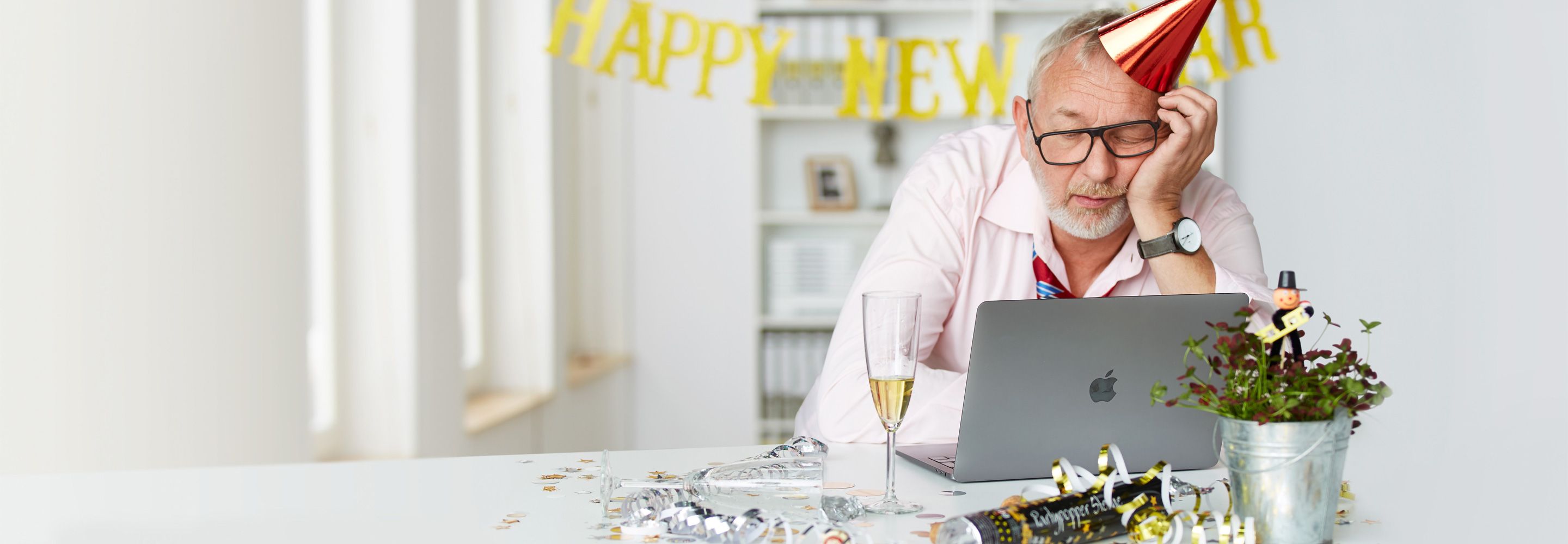 Een man met een feestmuts zit te dommelen voor een laptop. Op zijn bureau staat een glas champagne en de tafel is versierd voor oudejaarsavond. 