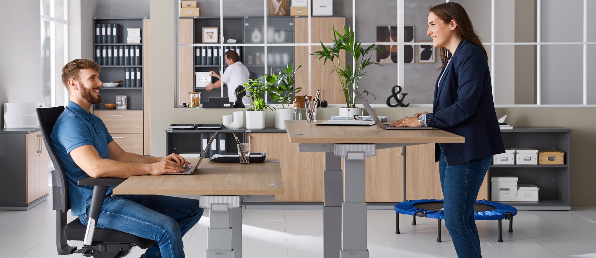 Ein Mann und eine Frau arbeiten im Sitzen und Stehen ergonomisch an ihren Schreibtischen