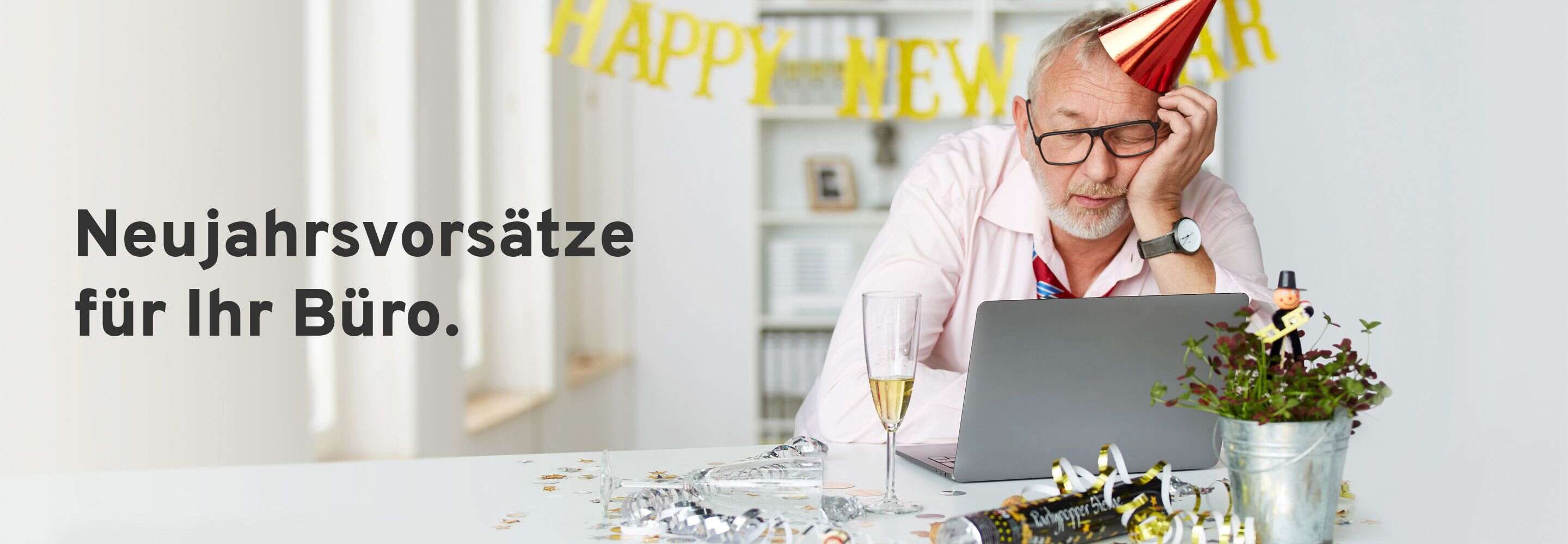 Ein Mann mit Partyhut döst vor einem Laptop. Auf seinem Schreibtisch steht ein Glas Sekt und der Tisch ist zu Silvester dekoriert. 
