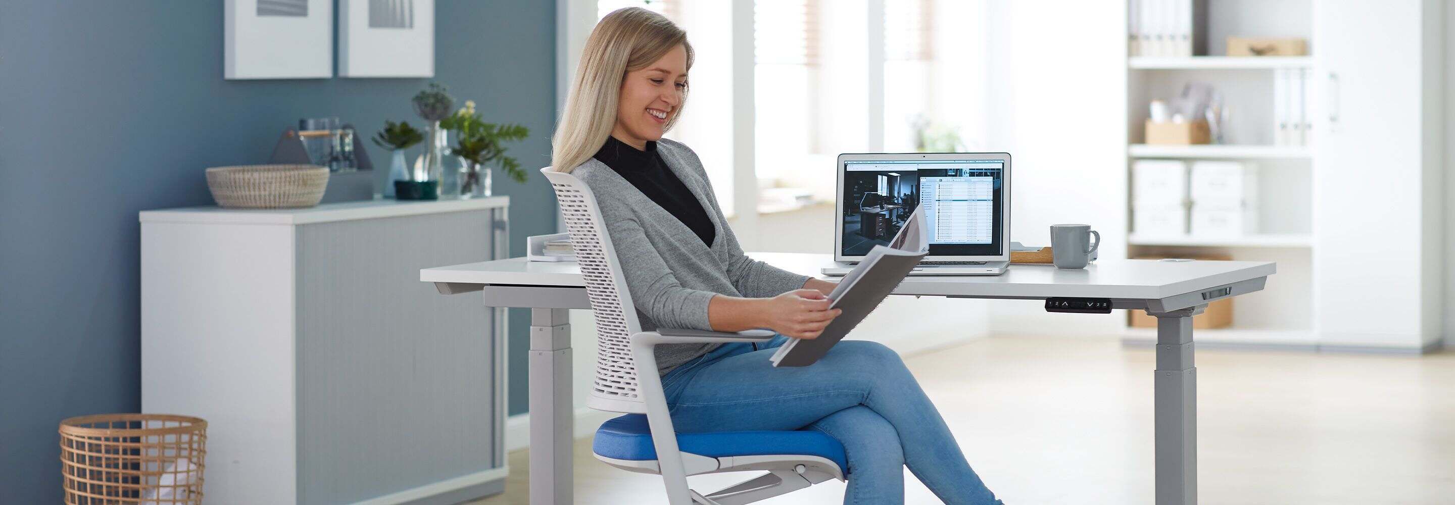 Een vrouw zit glimlachend achter een bureau en leest iets.