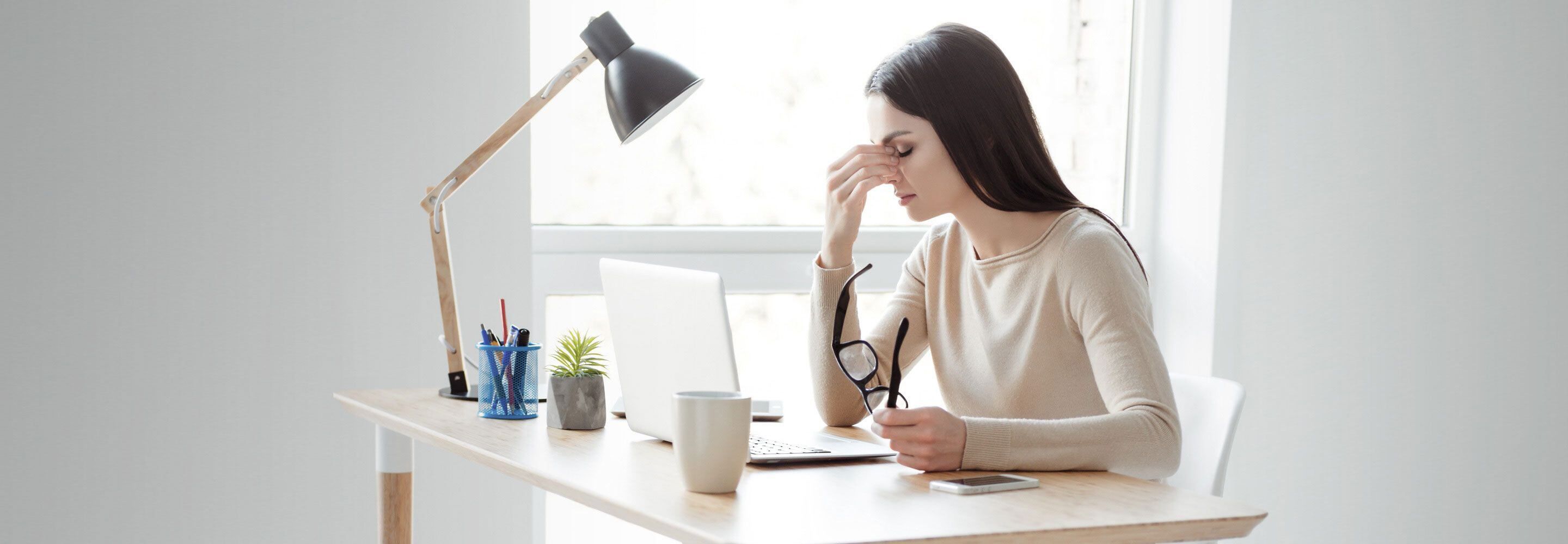 Een vrouw zit aan een bureau. Ze wrijft in haar ogen en houdt haar bril in haar hand. Voor haar staat een laptop.