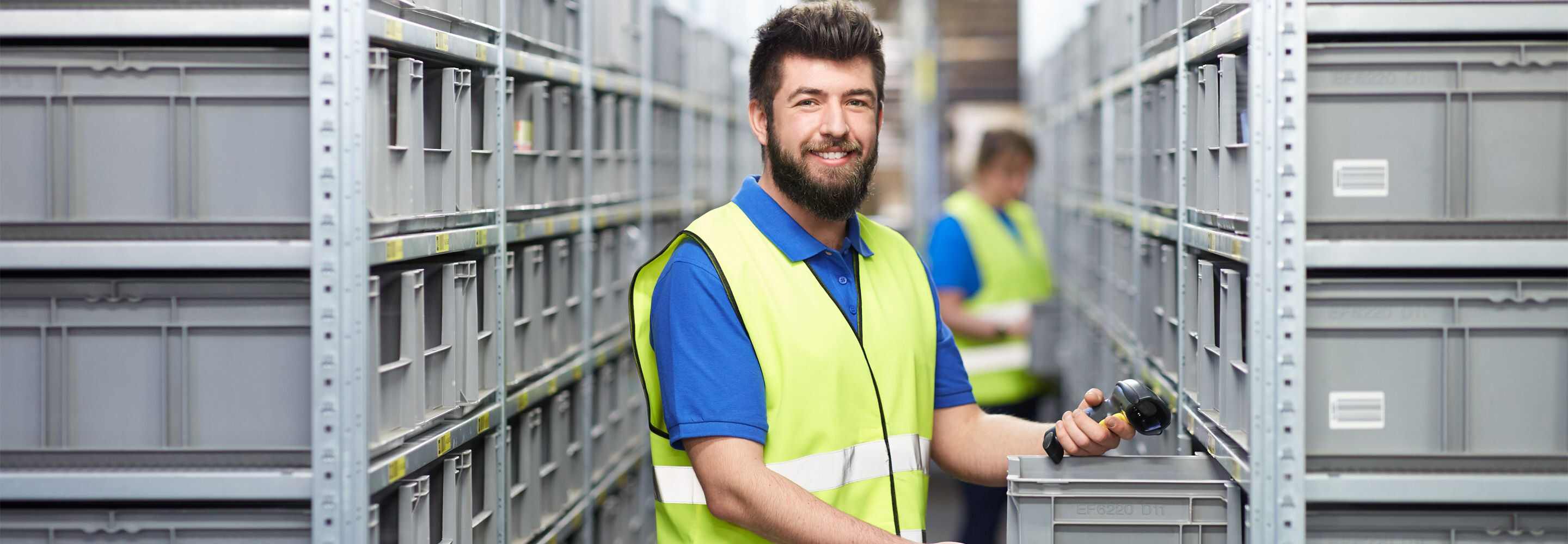 Een man met een high-visibility vest lacht in de camera en staat in een magazijn.