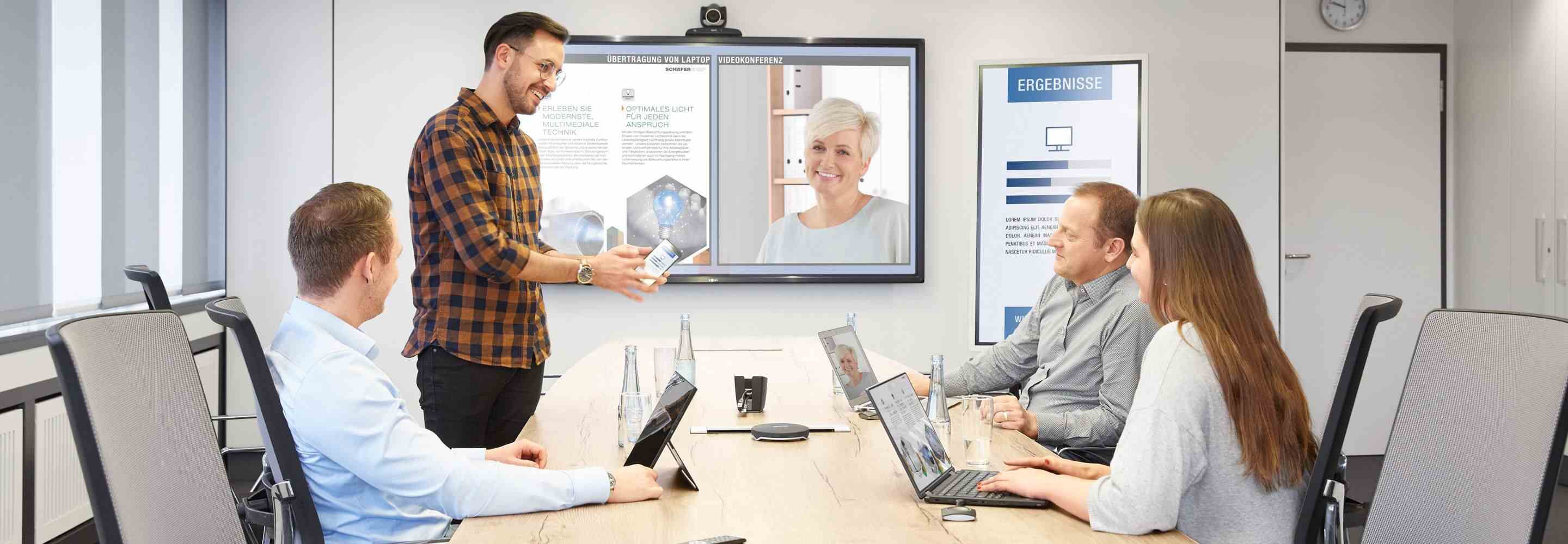 Drei Männer und eine Frau sitzen an einem Konferenztisch. Eine weitere Frau nimmt virtuell am Meeting Teil und ist auf einem großen Bildschirm zu sehen.