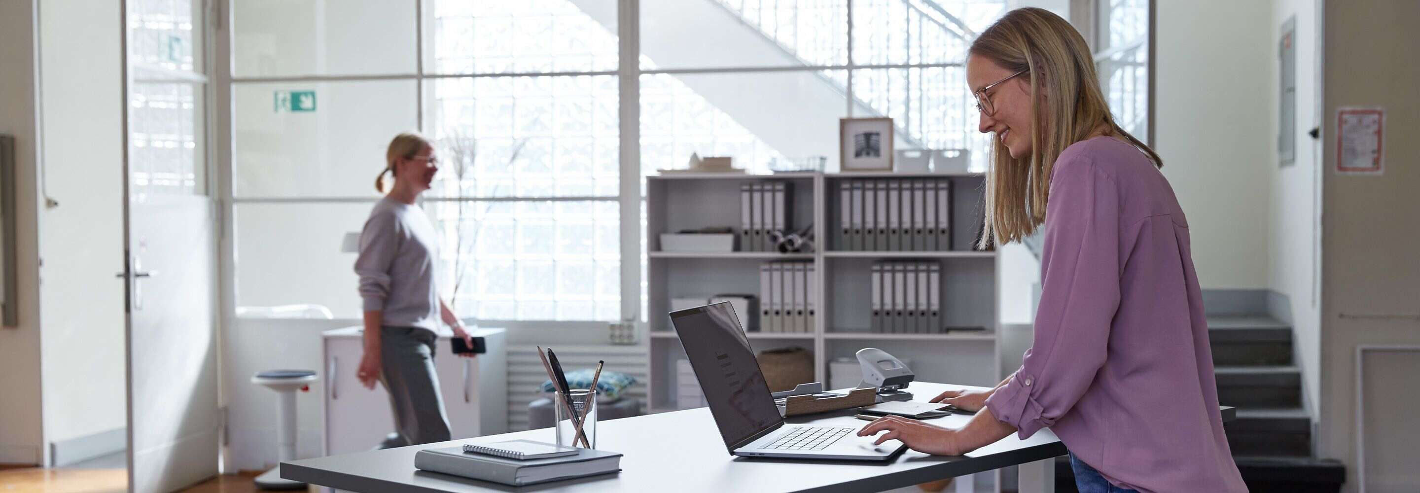 Eine Frau steht an einem Schreibtisch und arbeitet an einem Laptop. Eine andere Frau läuft im Hintergrund durch das Büro.