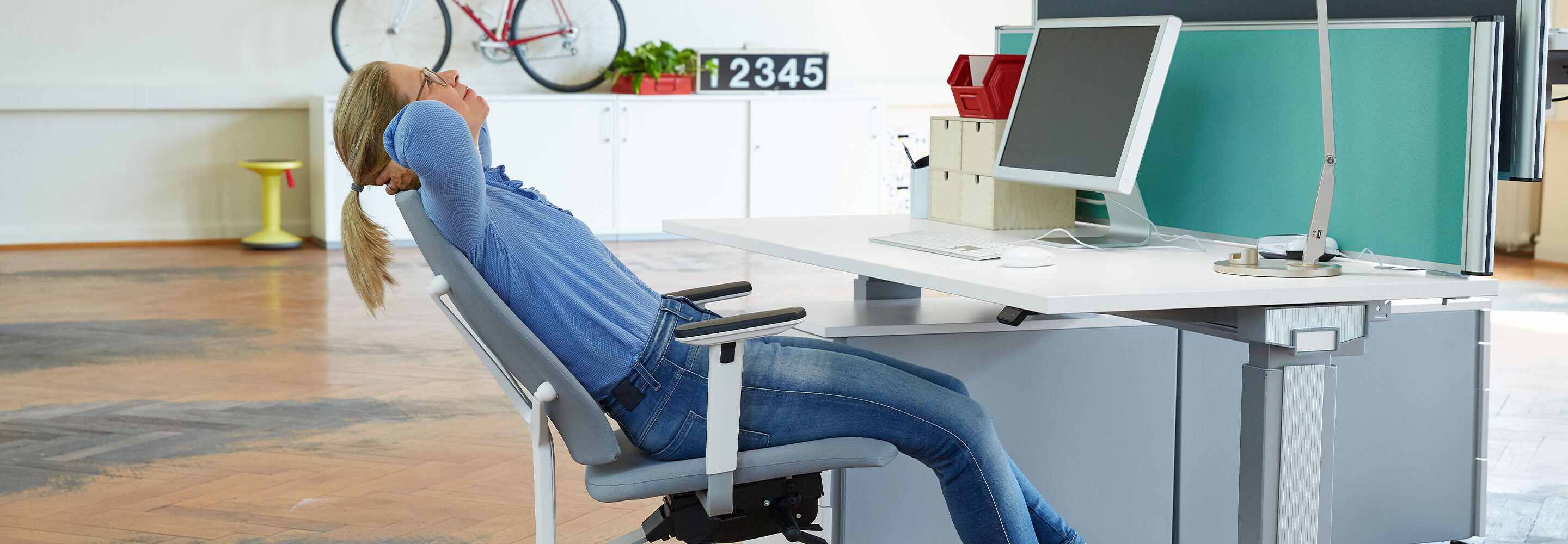 Une femme se penche en arrière sur sa chaise de bureau, les bras repliés derrière la nuque. 