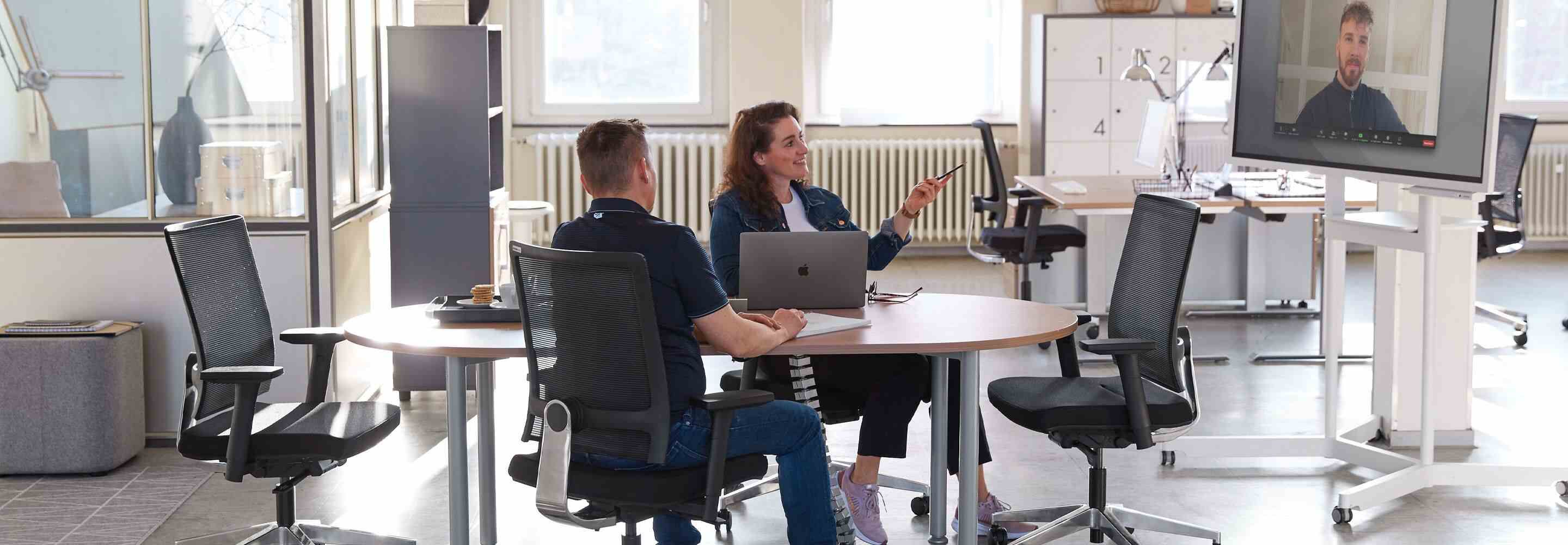 Een vrouw en een man zitten aan een vergadertafel. Een andere man is virtueel verbonden via een scherm.