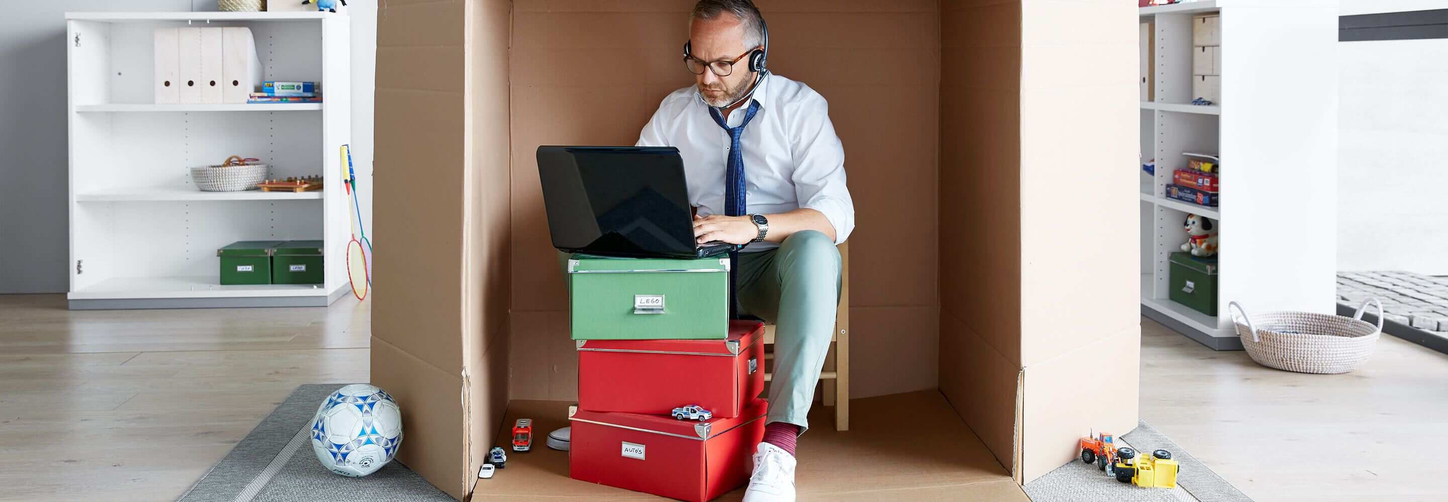 Een man zit in een papieren doos, die hij heeft omgebouwd tot werkplek, in de kamer van een kind.