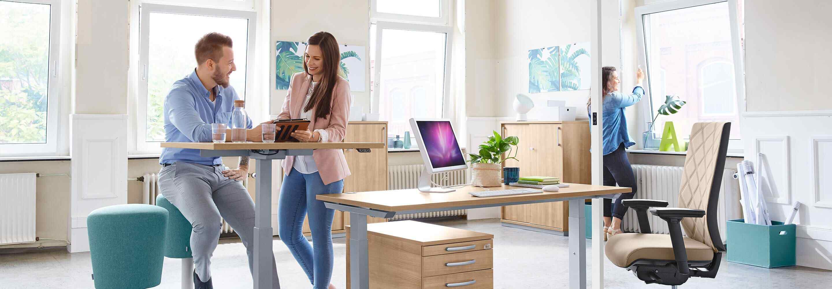 Een vrouw opent een raam in het kantoor. Een andere vrouw en een man staan aan het bureau en laten elkaar iets zien op een tablet.