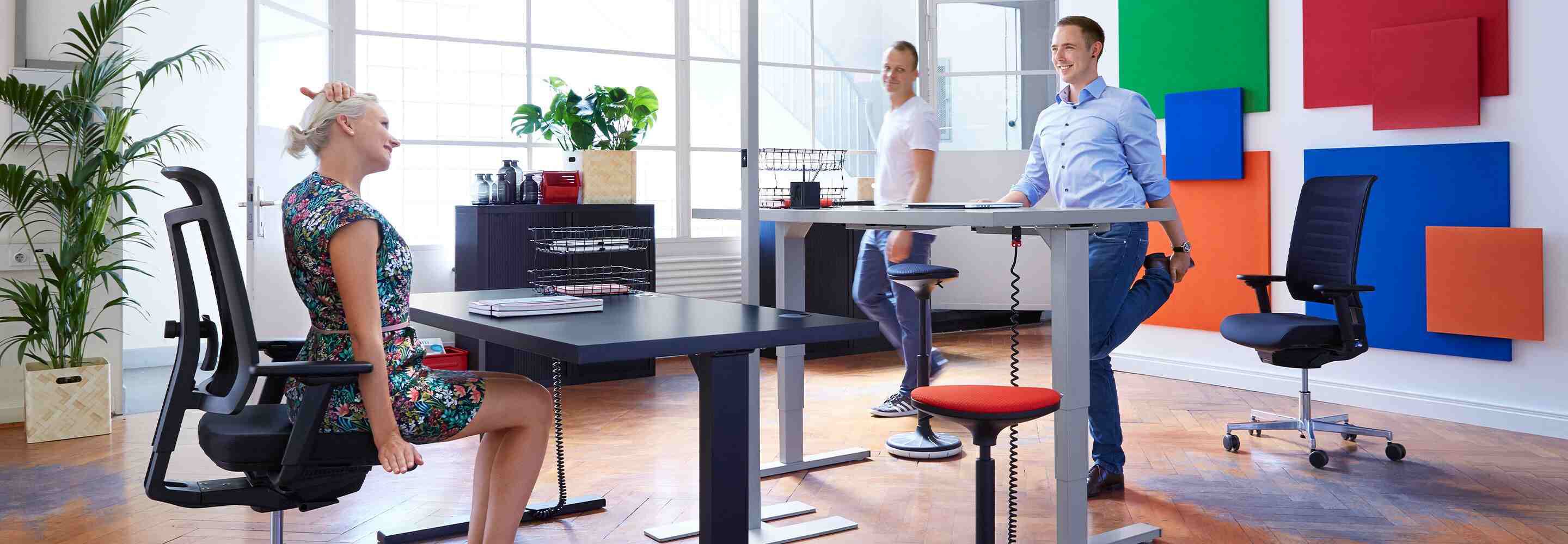 Eine Frau dehnt sich am Schreibtisch während zwei Männer sportliche Übungen im Stehen machen. Alle drei machen kurze Workouts. 