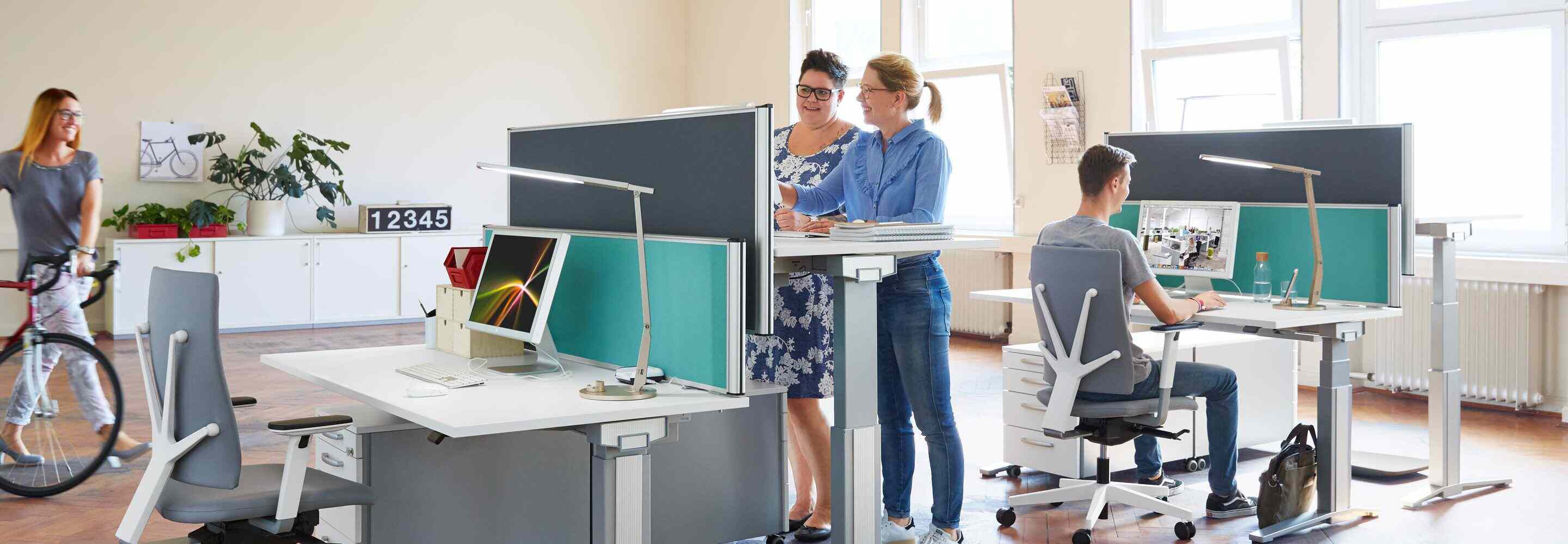Trois femmes et un homme dans un bureau paysager. Une femme pousse son vélo dans le bureau, les autres travaillent sur des ordinateurs. 
