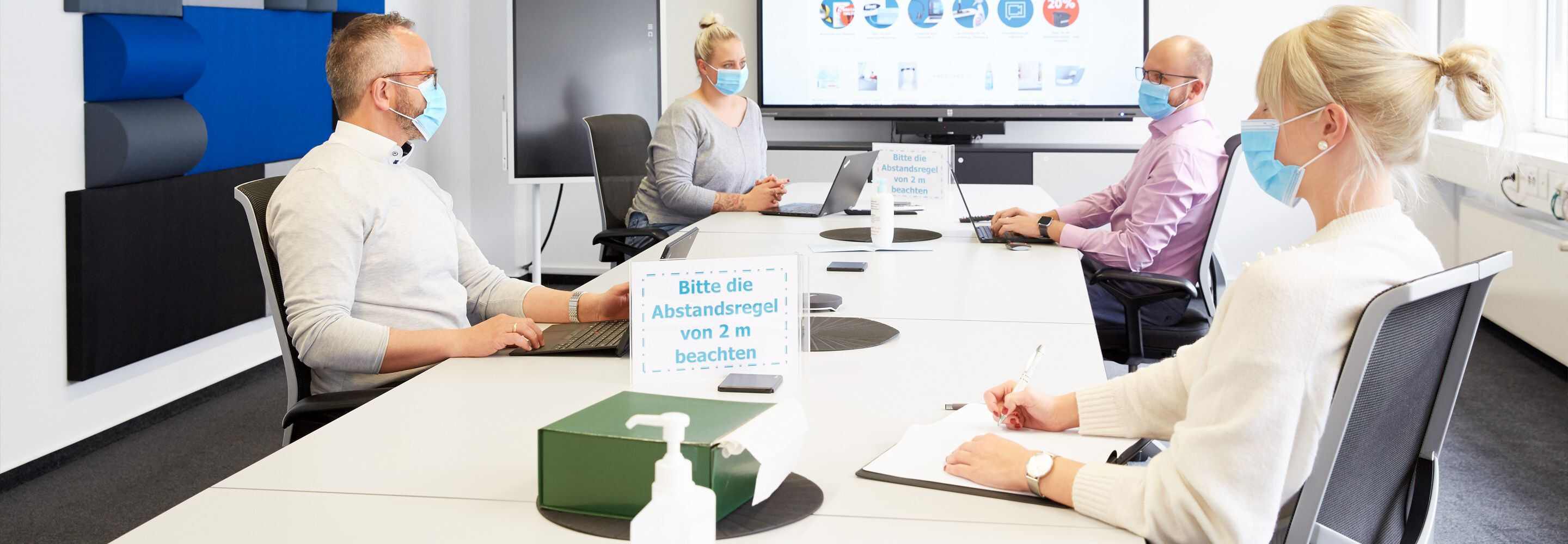 Vier mensen zitten met hun laptop aan een tafel. Allemaal hebben ze een mondkapje op.