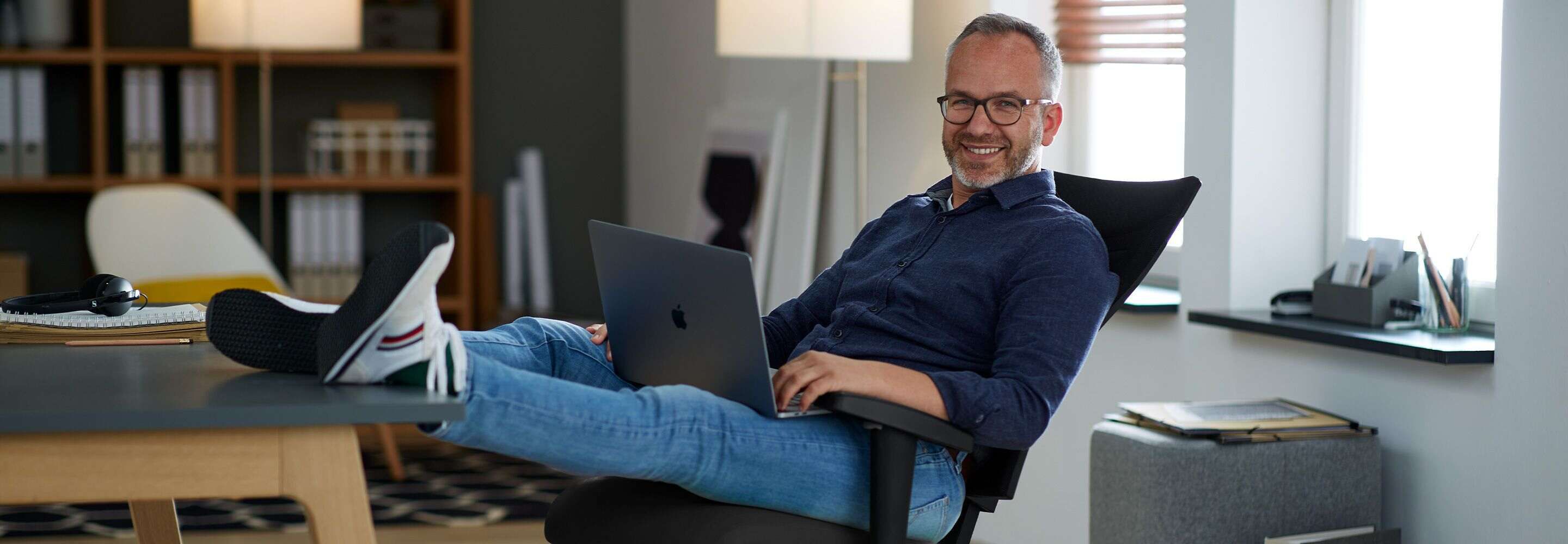 Een man heeft zijn voeten op zijn bureau gelegd en lacht naar de camera met een laptop op zijn schoot. 