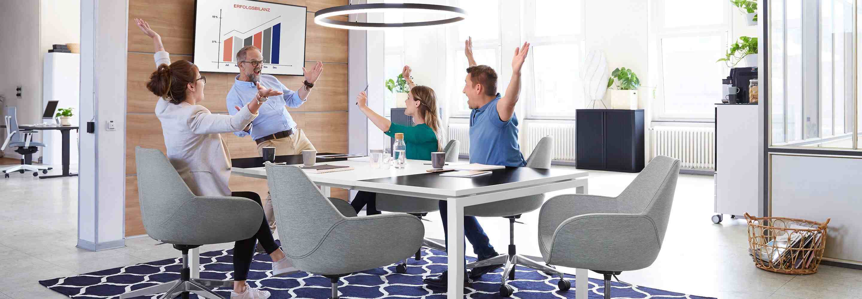 Vier Menschen sitzen an einem Konferenztisch im Büro und jubeln. Sie haben die Arme hochgerissen und freuen sich.