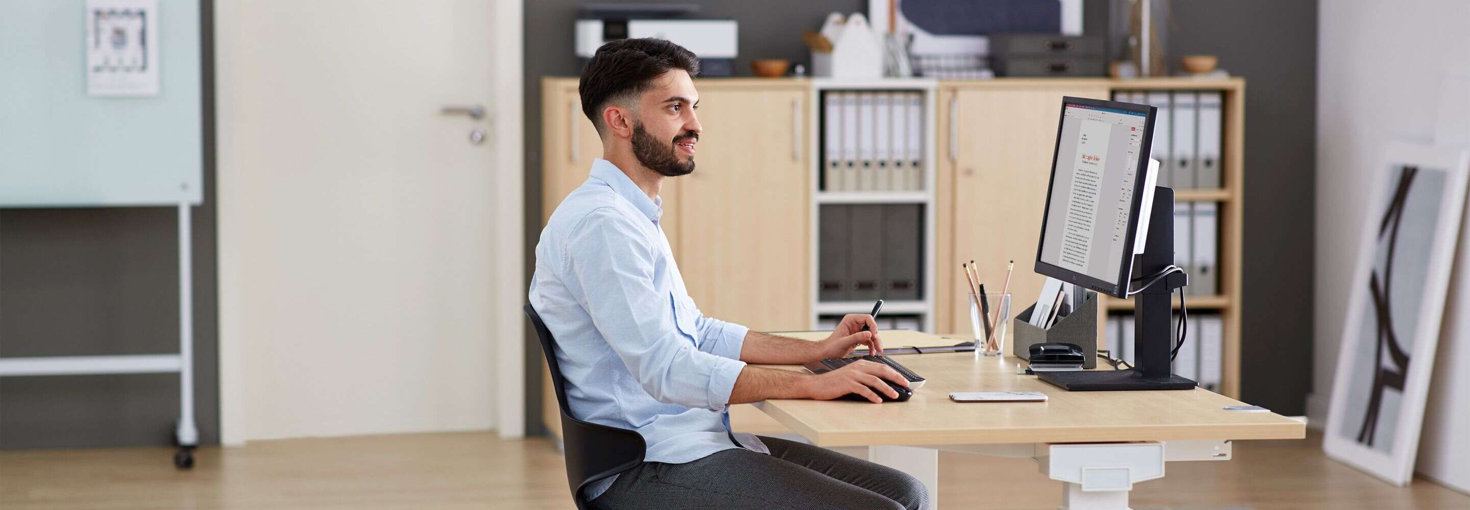 Mann sitzt lächelnd am Schreibtisch und klickt mit seiner Maus auf seinem Computerbildschirm.