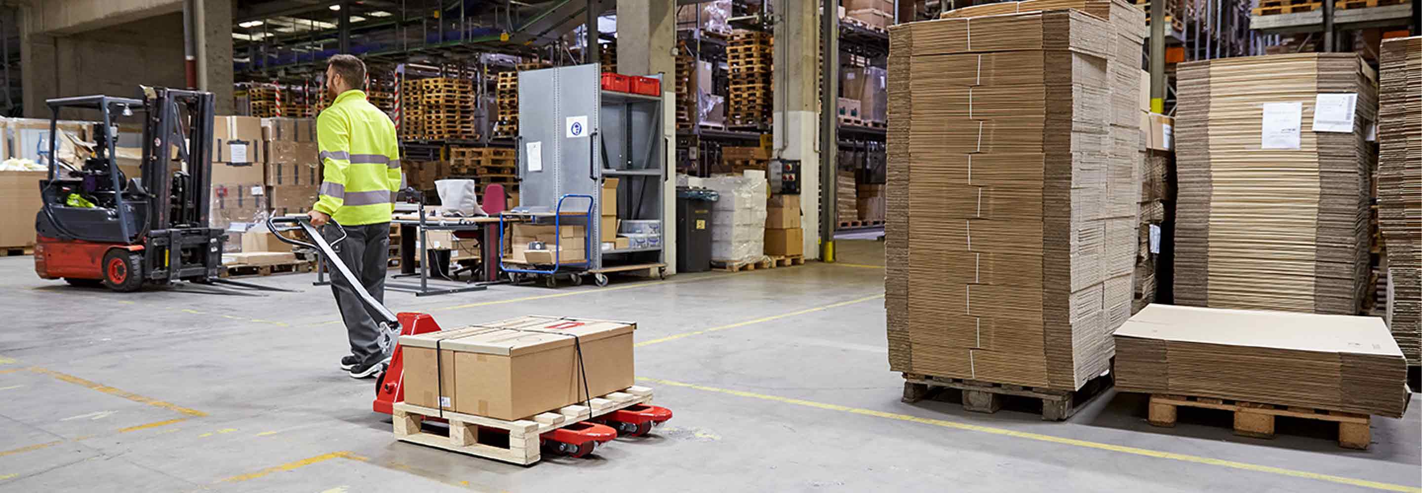 Een man draagt een high-visibility vest in een magazijn en trekt een pallettruck achter zich aan.