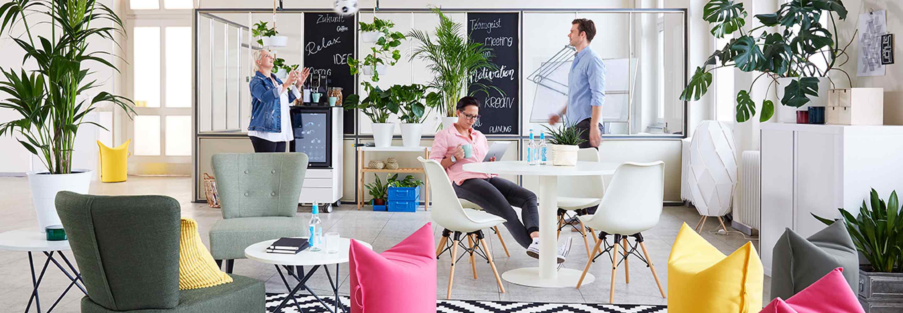 Ein bunt eingerichtetes Büro, im Vordergrund stehen Stühle, Sessel und Sitzsäcke. Im Hintergrund sieht man ein verglastes Büro und einen Kaffeeautomaten, an dem sich zwei Frauen begegnen. Im Büro arbeitet ein Mann.