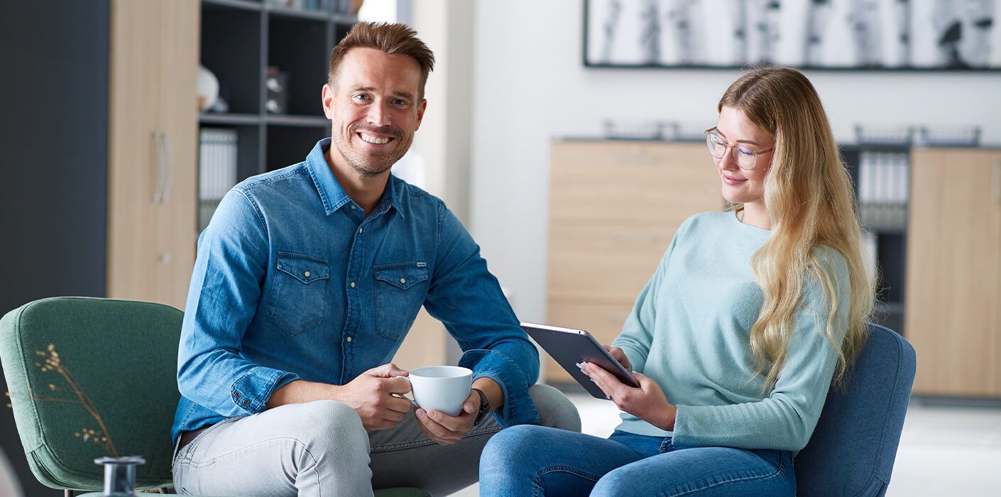 Eine Person mit einer Kaffetasse in der Hand sitzt einer Person mit einem Tablet in der Hand gegenueber