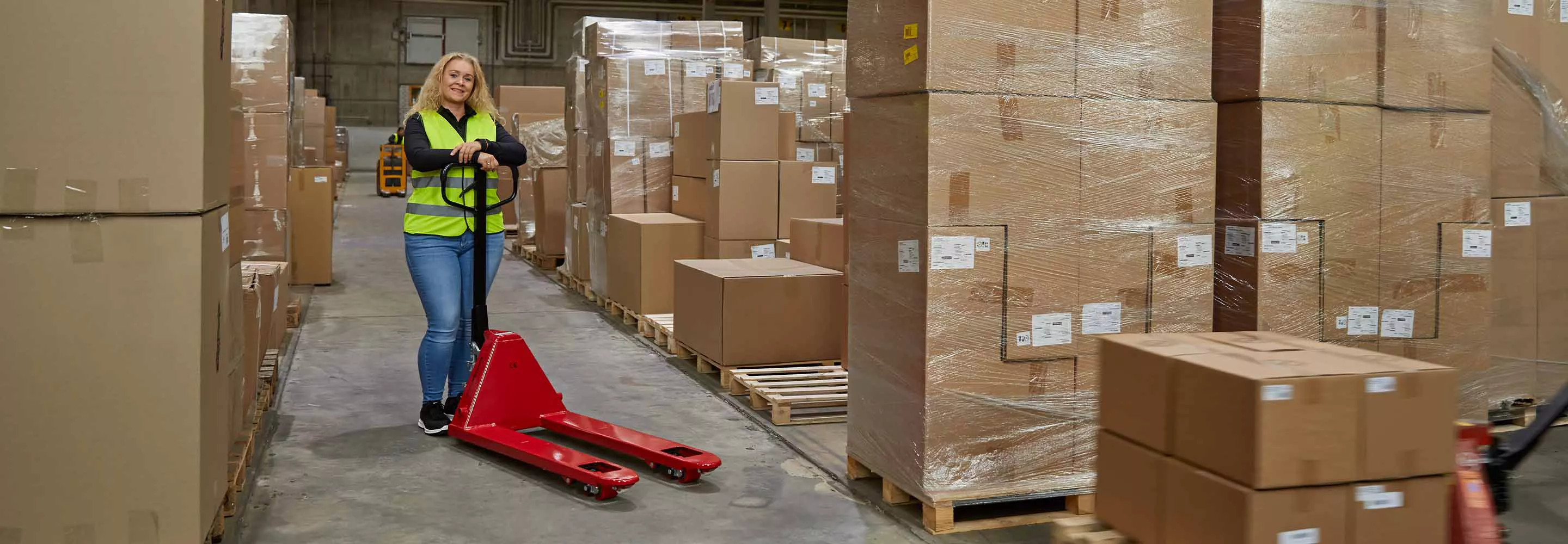 Une femme vêtue d'un gilet de sécurité jaune se tient debout dans un entrepôt au milieu de cartons et s'appuie sur le timon d'un chariot élévateur rouge.