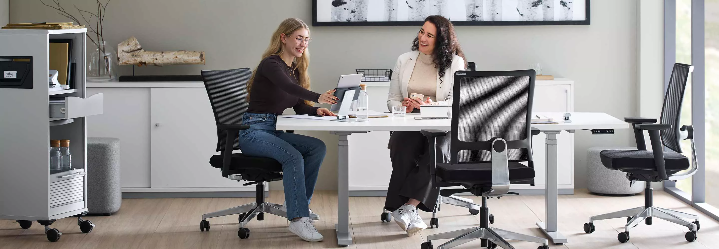 Zwei Frauen sitzen in einem Büro an einem weißen höhenverstellbaren Schreibtisch. Eine Frau hält ein Tablet und zeigt ihrer Kollegin etwas darauf. 