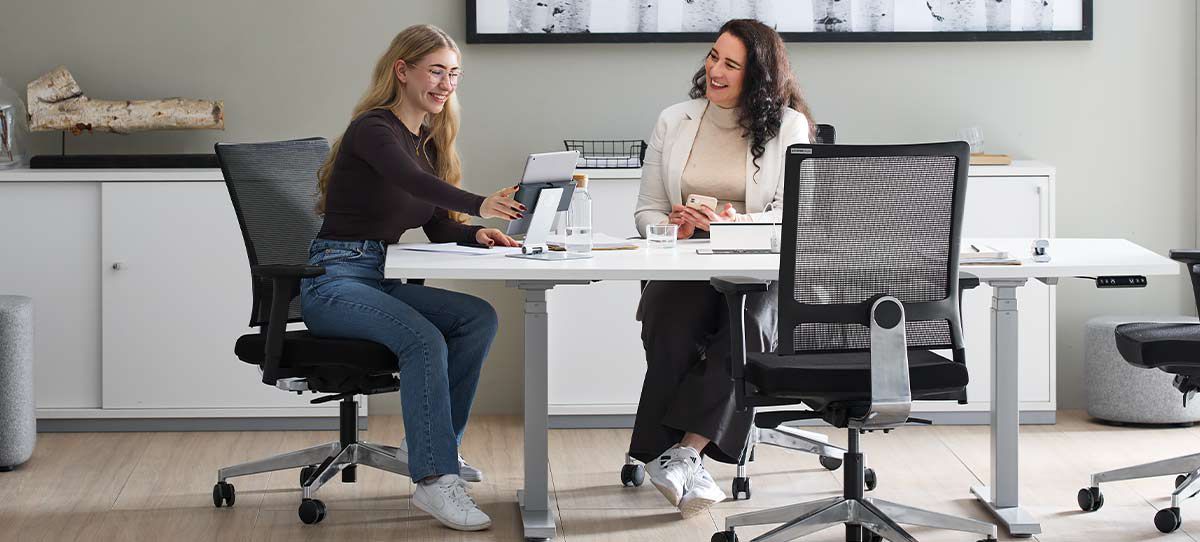 Zwei Kolleginnen von Schäfer Shop sitzen in ihrem modernen Büro. Sie betrachten ein Tablet und lachen.