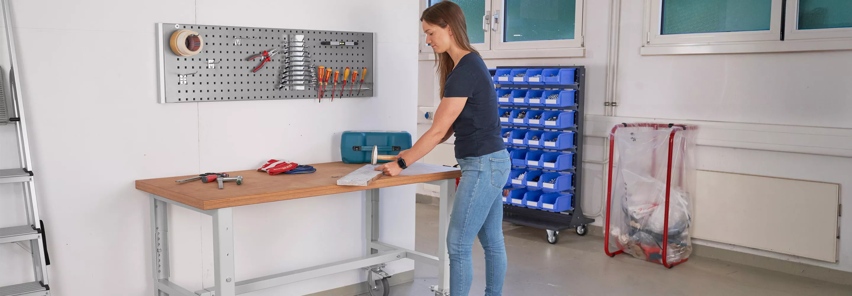 Frau arbeitet an einer mobilen Werkbank mit Holzplatte in einer Werkstatt, umgeben von Werkzeugen und Aufbewahrungssystemen.