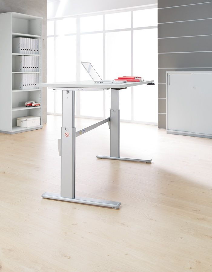 Höhenverstellbarer Stehschreibtisch in einem modernen Büro mit hellem Holzfußboden und großer Fensterfront, auf dem ein aufgeklappter Laptop und rote Ordner liegen.