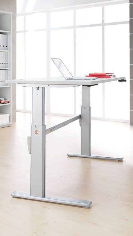 Höhenverstellbarer Stehschreibtisch in einem modernen Büro mit hellem Holzfußboden und großer Fensterfront, auf dem ein aufgeklappter Laptop und rote Ordner liegen.