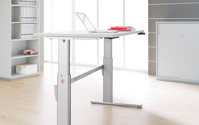 Höhenverstellbarer Stehschreibtisch in einem modernen Büro mit hellem Holzfußboden und großer Fensterfront, auf dem ein aufgeklappter Laptop und rote Ordner liegen.