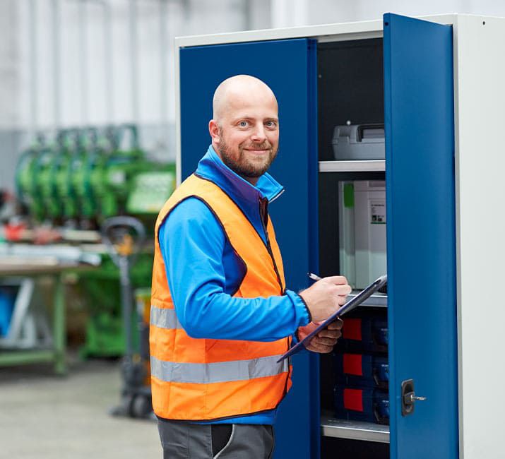 Mitarbeiter kontrolliert Inhalt eines blauen Schranksystems und notiert Daten auf Klemmbrett in Produktionsumgebung