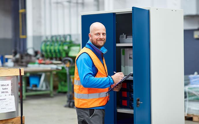 Mitarbeiter kontrolliert Inhalt eines blauen Schranksystems und notiert Daten auf Klemmbrett in Produktionsumgebung