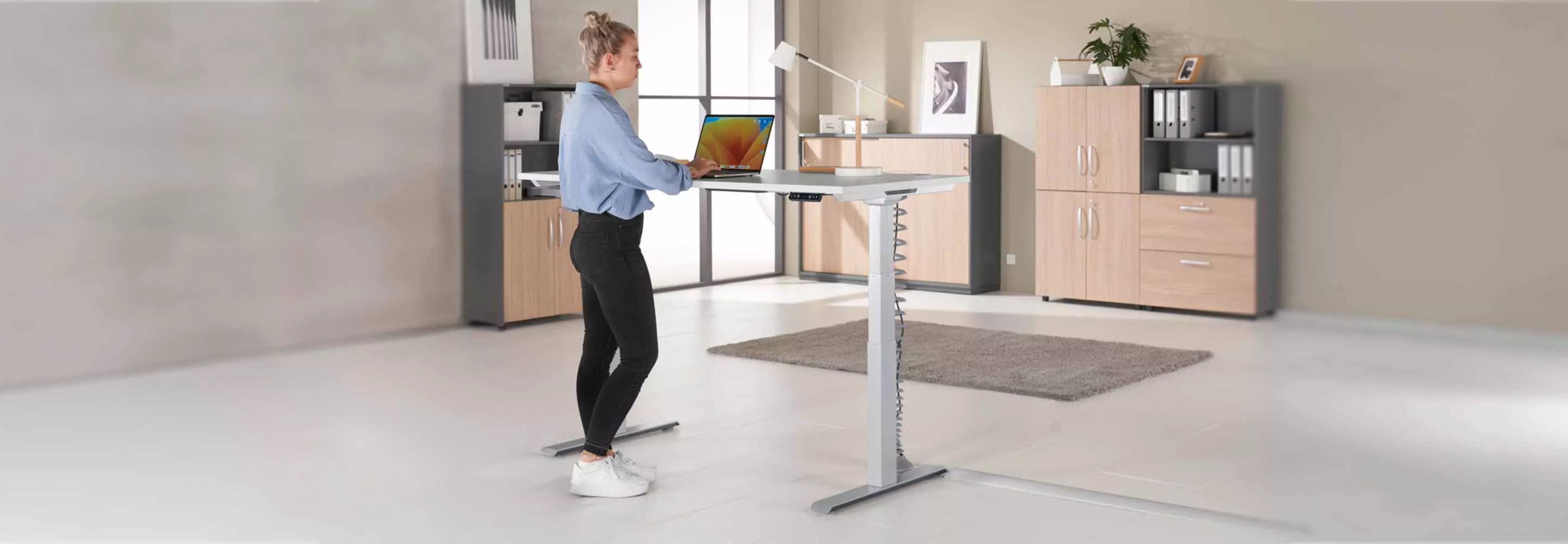Frau sitzt an höhenverstellbarem Schreibtisch und arbeitet am Laptop. Büroeinrichtung mit Schränken.