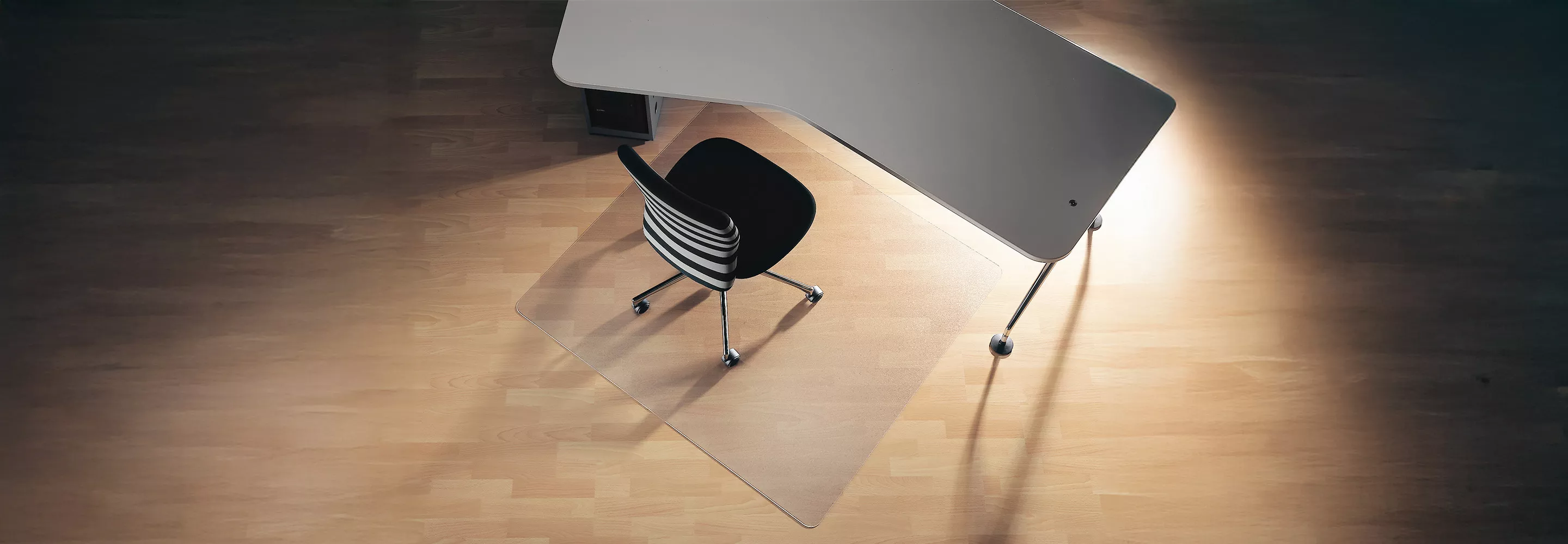 Blick von oben auf Schreibtisch mit Stuhl und Bodenschutzmatte. Der Boden ist helles Holz. Der Stuhl ist schwarz mit weißen Streifen.