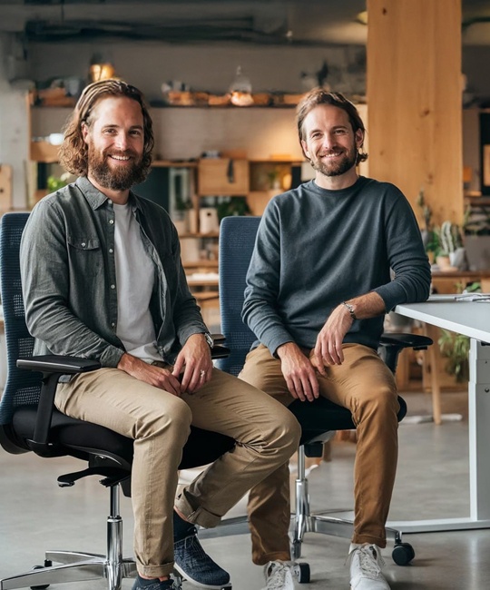 Zwei Kollegen, die in einem modernen Büro nebeneinander auf Bürostühlen sitzen und in die Kamera lächeln