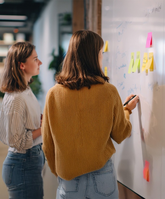 Zwei junge Frauen stehen in einem Büro vor einem Whiteboard mit Notizen.