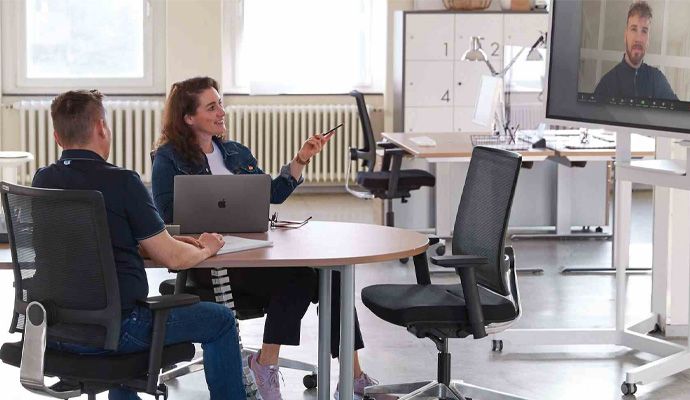 Eine Frau und ein Mann sitzen an einem Besprechungstisch. Ein weiterer Mann ist virtuell über einen Bildschirm zugeschaltet.