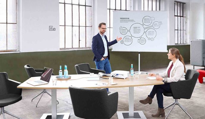 Eine Frau sitzt an einem Konferenztisch und hört einem Mann zu, der etwas an einer Leinwand präsentiert.