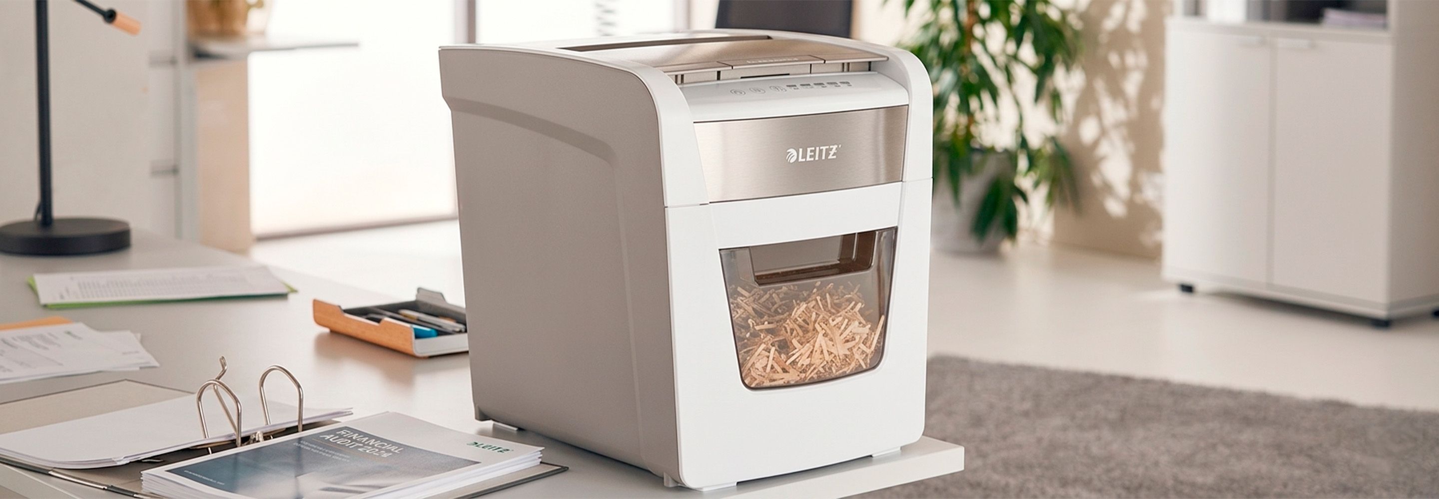 Ein Leitz-Aktenvernichter auf einem modernen Schreibtisch in einer Büroumgebung. Um ihn herum liegen Akten.