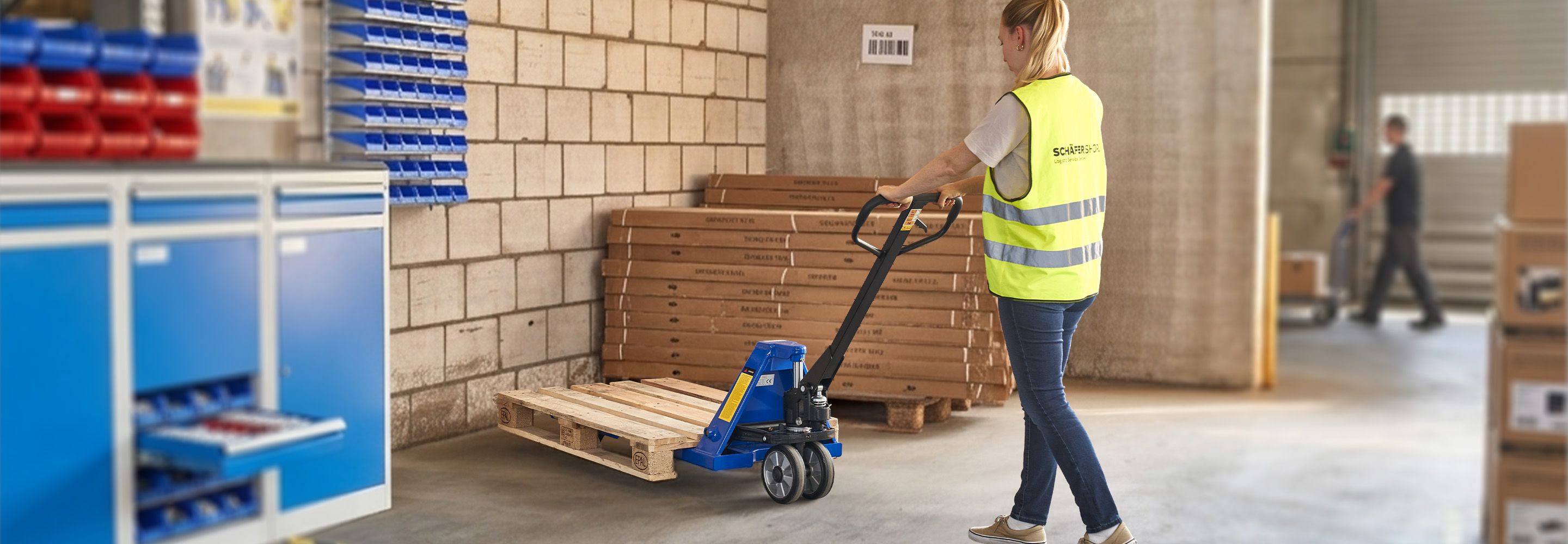 Eine Mitarbeiterin in Warnweste zieht eine leere Europalette mit einem blauen Hubwagen durch das Lager