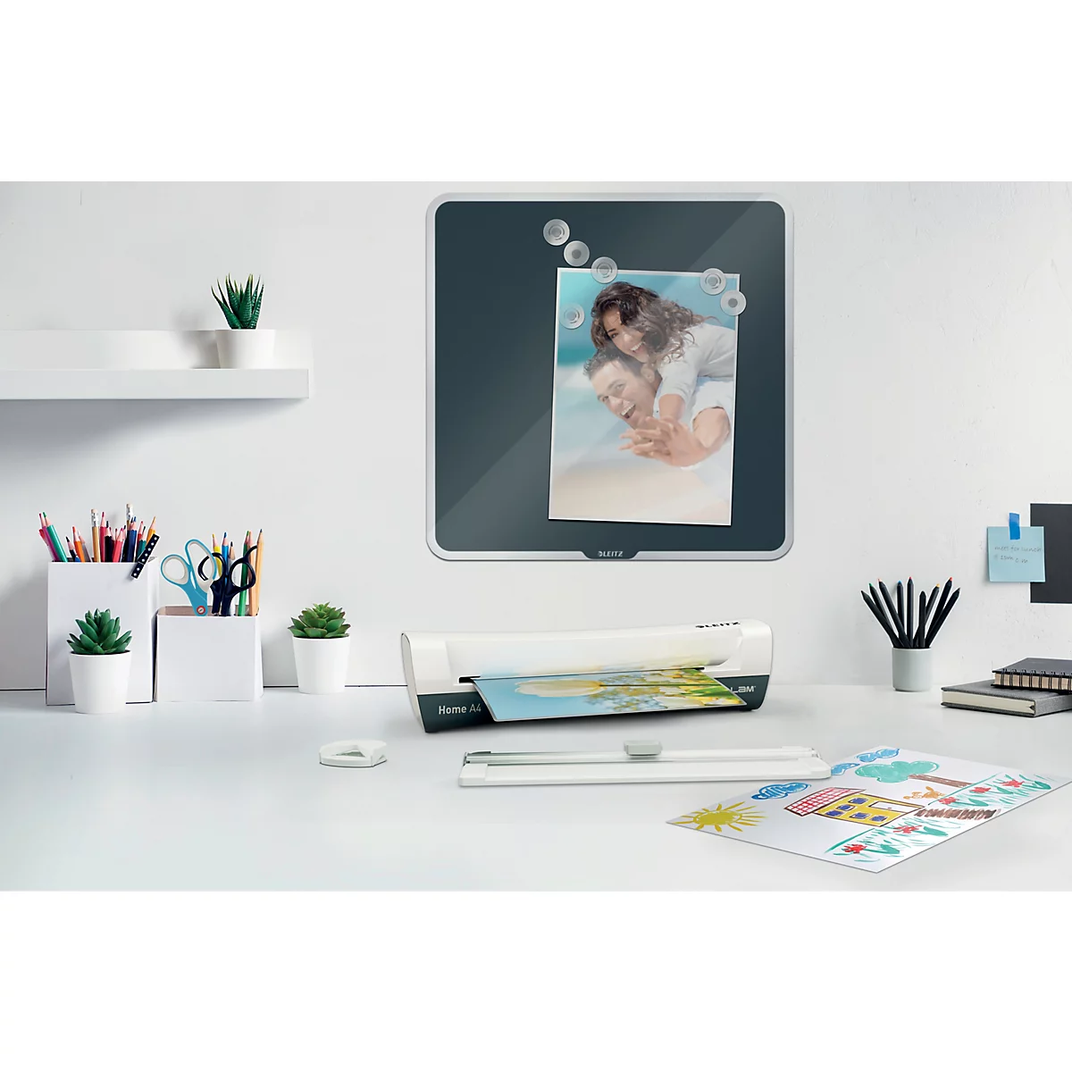 Bureau met lamineermachine, magneetbord met foto, planten en pennen.
