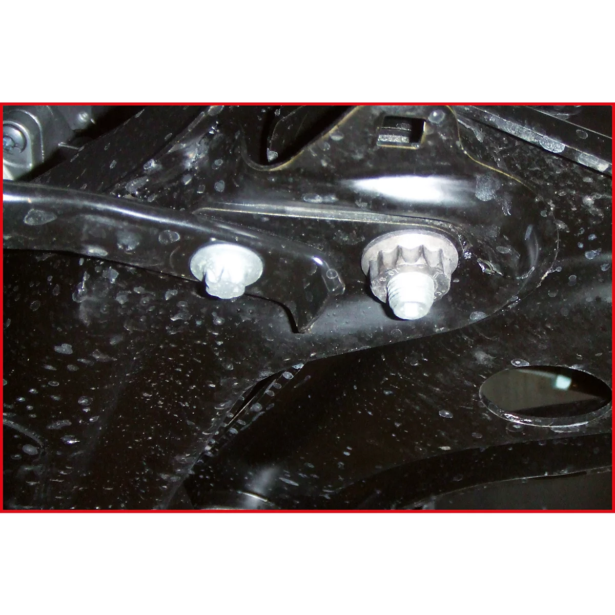 Nahaufnahme von zwei Schrauben, die an einem schwarzen Metallrahmen befestigt sind. Das Metall hat kleine Unebenheiten.