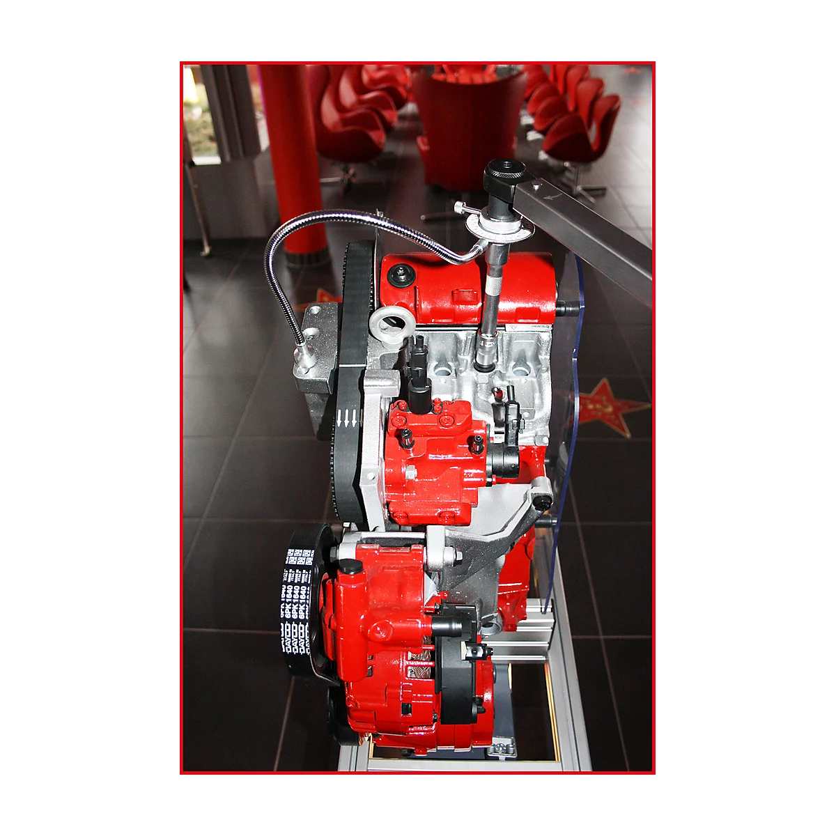 Motor im Schnitt. Rote und silberne Komponenten sind erkennbar. Der Hintergrund zeigt rote Stühle und einen Stern.